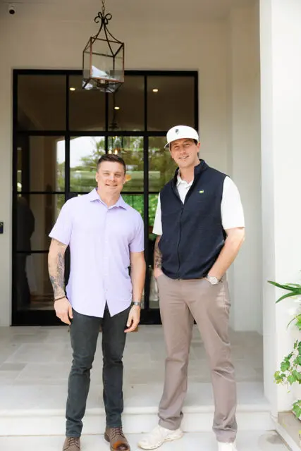 Two men standing on a porch. The man on the left wears a lavender shirt and dark jeans.