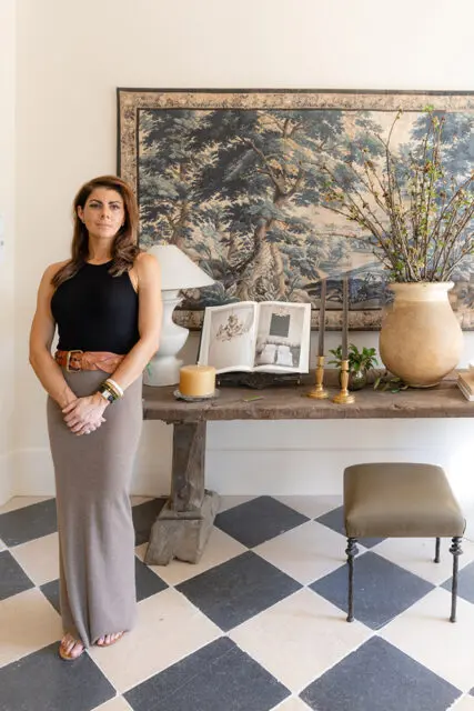 Woman in taupe dress and black top standing by console table with tapestry