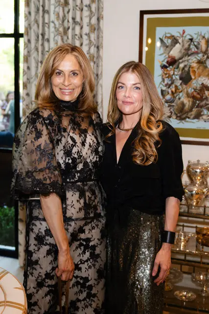 Two women posing indoors. One wears a black lace dress, the other a black shirt and sequined skirt.