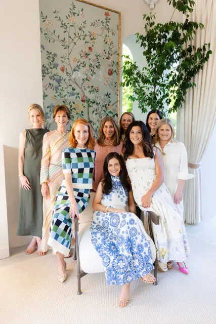 Group of eight women posing indoors, with a floral mural and large plant behind them.