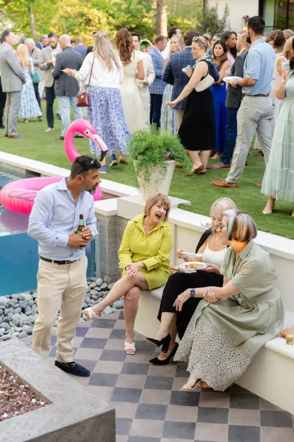 People gathered around a pool with a flamingo float, some sitting on a bench and talking.
