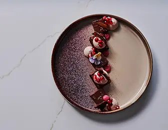 Dessert on a speckled plate with chocolate, cream, pomegranate seeds, and edible flowers.