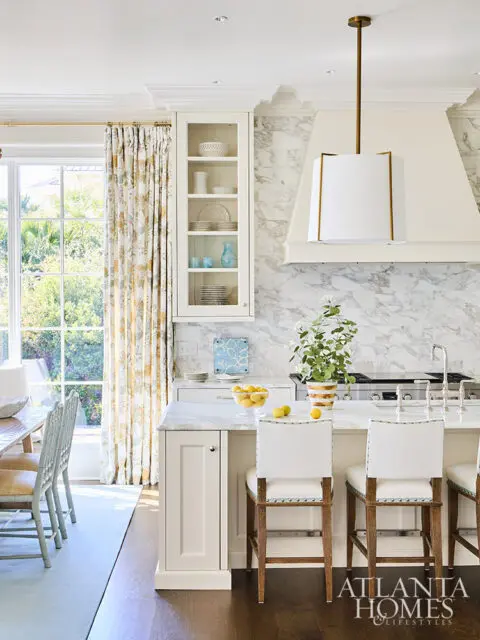 White kitchen island with marble countertop, white bar stools, and marble backsplash.