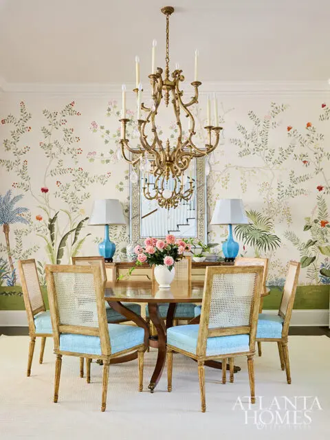Dining room with floral wallpaper, cane-back chairs with blue cushions, and ornate chandelier.