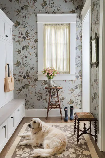 Mudroom with floral wallpaper, white built-in bench and cabinets, and a golden retriever on a rug.