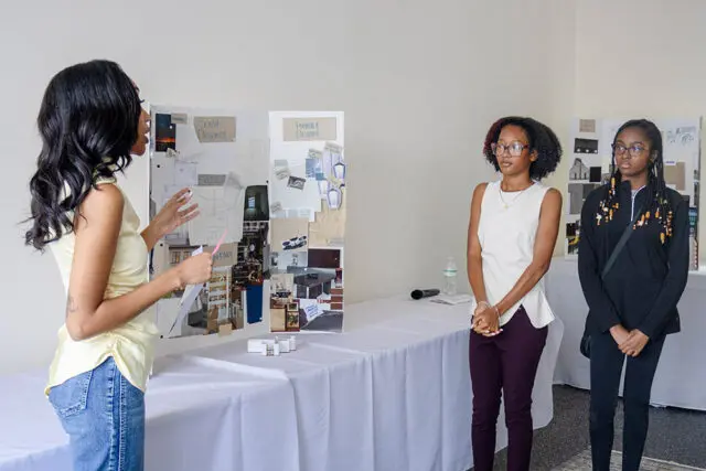 Three young women stand near a presentation board with design collages and architectural drawings.