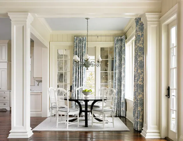 Dining room with white chairs around a dark round table, blue floral curtains, and white paneled