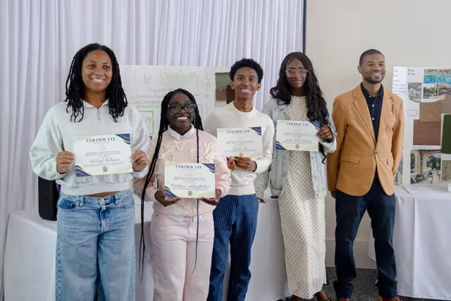 Five people stand holding certificates. A display board behind them has text that reads 'The