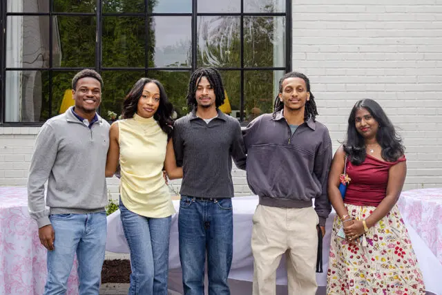 Five people stand in front of a white brick wall and a large window.