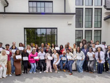 Group of students and volunteers holding certificates in front of a white brick building.