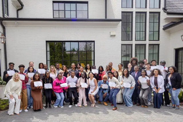 Group of students and volunteers holding certificates in front of a white brick building.