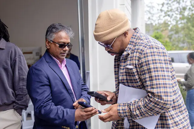 Man in blue suit and pink shirt shows phone to man in plaid shirt and beanie
