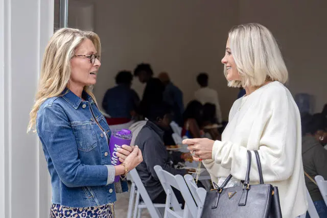 Two women talking, one holds a purple tumbler with 'MOM' written on it.