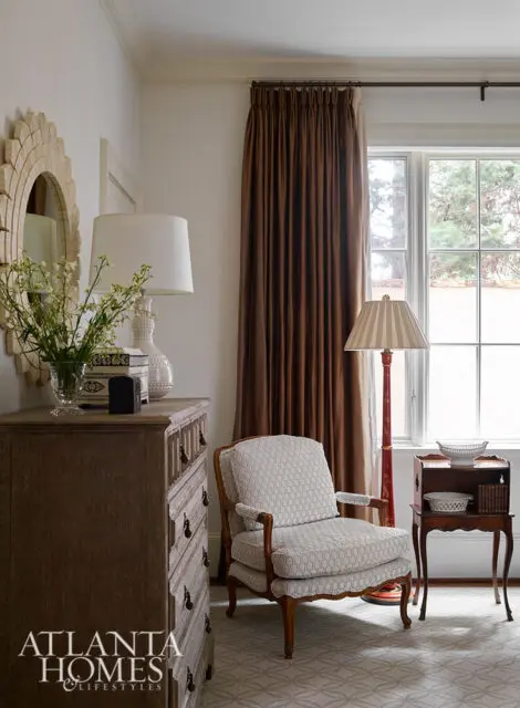 Light-filled bedroom with a light wood dresser, upholstered armchair, and floor-to-ceiling brown curtains.
