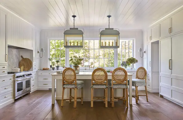White kitchen with wood floors, island with rattan stools, and large windows overlooking greenery.