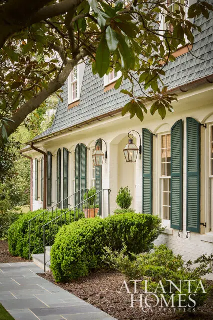 White brick house with dark green shutters, arched entryway, and gas lanterns. Slate walkway and manicured boxwood bushes.