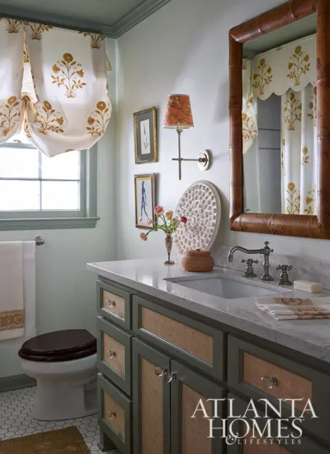 Powder room with green vanity, marble countertop, floral curtains, and bamboo mirror.