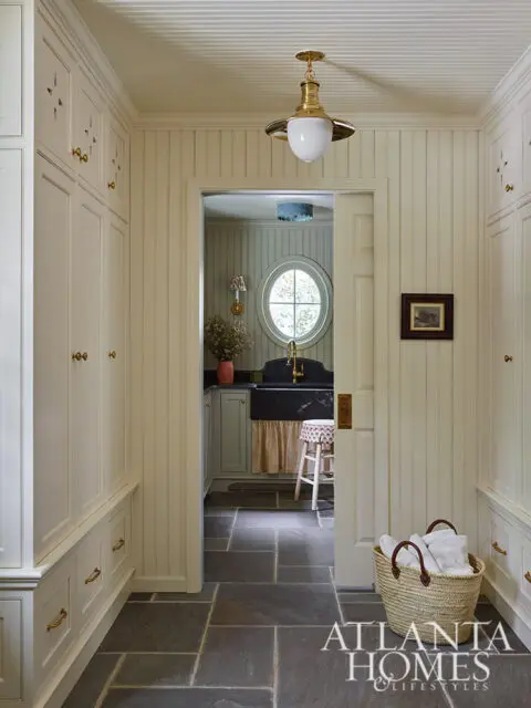 Cream-colored beadboard walls and custom cabinetry line a hallway with slate tile flooring. A doorway reveals a laundry room with an oval window.