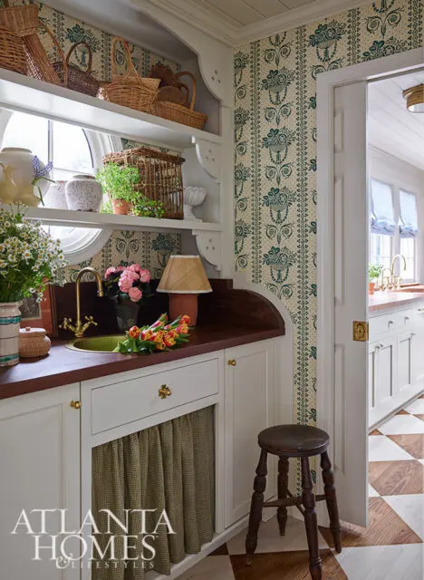 Cream and green floral wallpaper adorns a butler's pantry with white cabinetry, a brass sink, and a wooden stool.