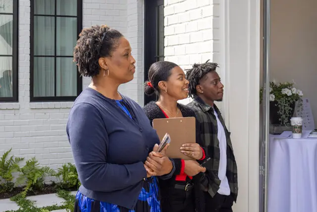 Three people standing outside a white brick building. The person in front is a woman wearing a navy
