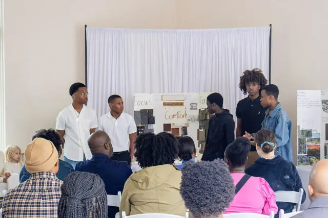 Group of young men presenting at a board with fabric swatches and design sketches.
