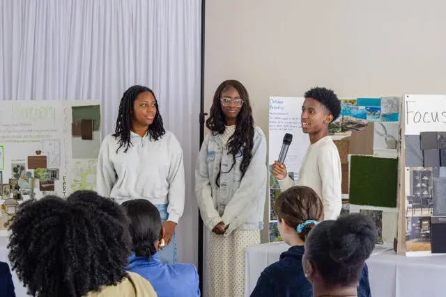 Three students presenting design boards, one holding a microphone.