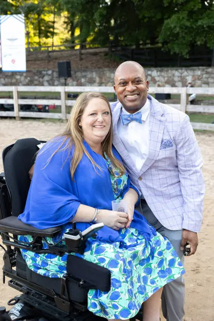 Woman in wheelchair wearing blue shawl and floral dress, man in checkered suit and bow tie