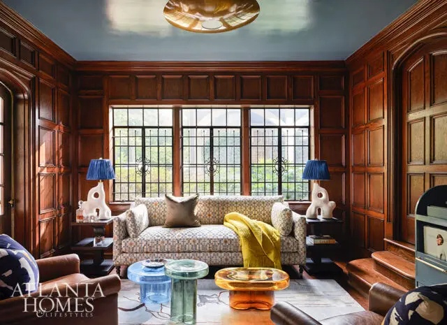 Living room with wood paneled walls, blue ceiling, patterned sofa, and colorful glass tables.