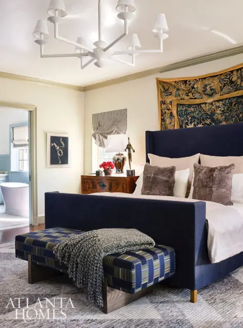 Bedroom with navy upholstered bed, patterned bench, tapestry, and white chandelier.