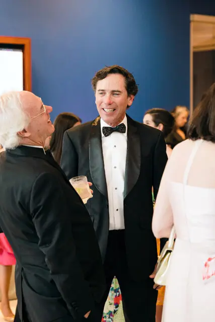 Man in a tuxedo smiles while holding a drink, talking to a man in a suit.