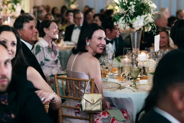 Woman in a pink dress smiling, seated at a formal dinner table with floral arrangements and place