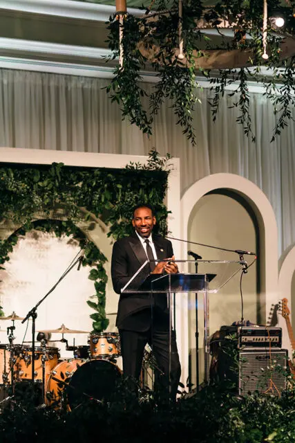 Man in tuxedo speaking at podium with drum set and guitar on stage