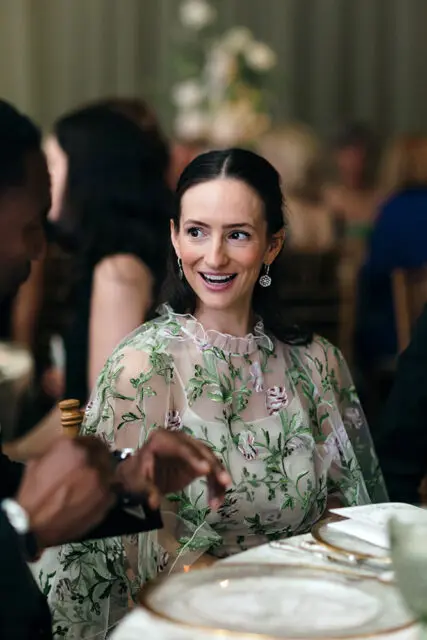 Woman in a sheer floral dress smiles at a table setting.