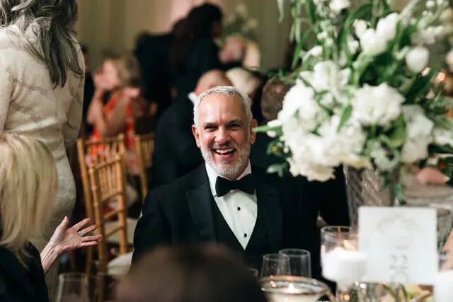 Man in tuxedo smiling, seated at a table with white floral centerpiece.