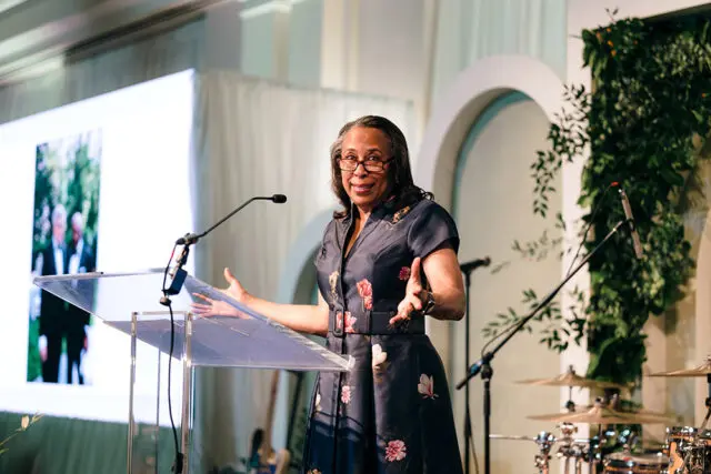 Woman in floral dress speaking at a podium with a screen behind her.