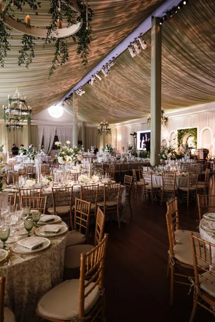 Banquet hall with draped ceiling, chandeliers, round tables set for dining, and bamboo chairs.