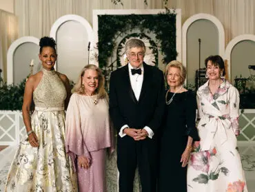 Five people in formal attire pose for a photo in front of a decorated backdrop.