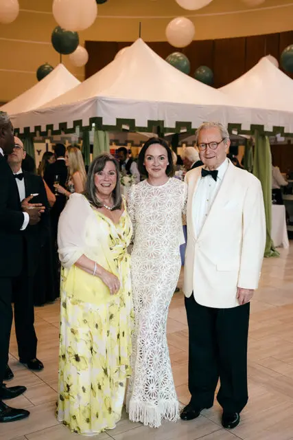 Three people posing in front of white tents and balloons at an event.