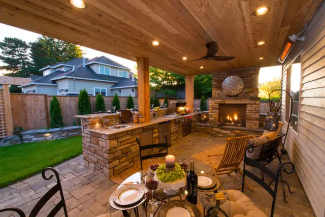 Covered outdoor kitchen with stone island, grill, fireplace, and dining table.