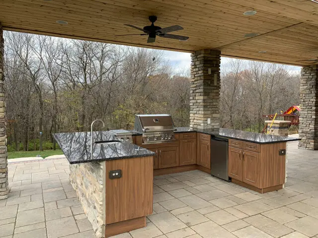 Outdoor kitchen with granite countertops, wood cabinetry, and stone pillars overlooking a wooded