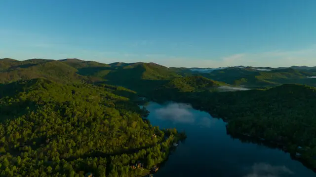 Aerial view of a lake surrounded by green, tree-covered mountains under a clear blue sky.
