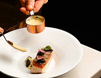 Chef garnishing a dish with sauce from a copper measuring cup.