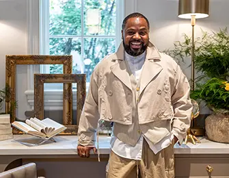 Man in tan jacket and pants smiles in a room with decorative frames and plants.