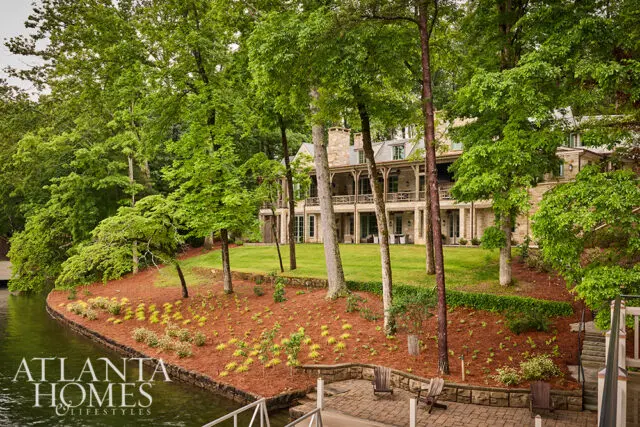 Stone mansion with multiple balconies overlooking a lake, surrounded by lush green trees.