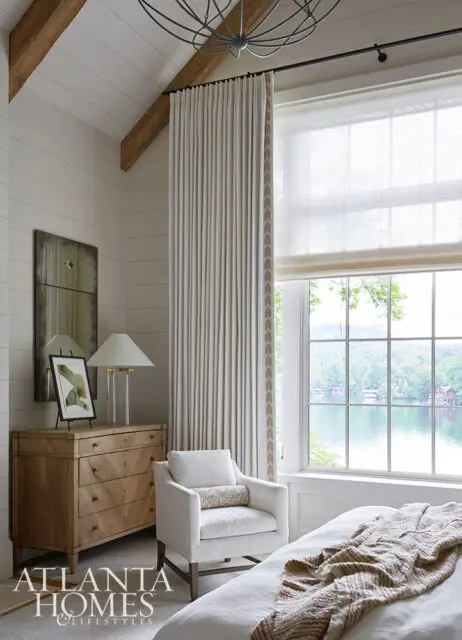 Bedroom with shiplap walls, wooden dresser, armchair, and large window overlooking a lake