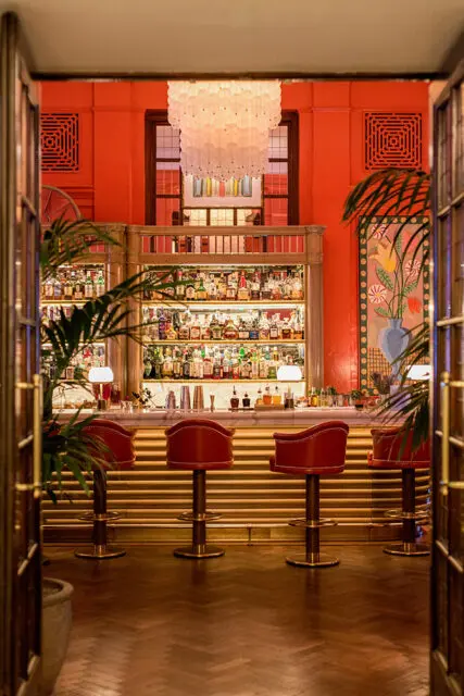 Bar with red leather stools, marble counter, and shelves of liquor bottles.