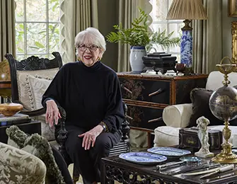 Elderly woman with white hair wearing glasses and a black sweater sits in an ornate chair in a