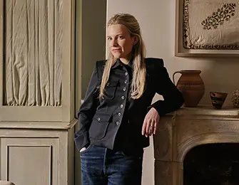 Woman with blonde hair wearing a black jacket and jeans, leaning against a fireplace mantel.