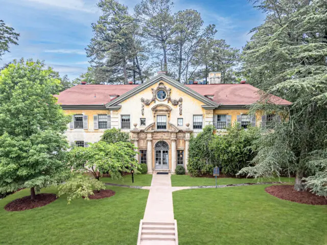 Yellow stucco mansion with a clock tower, surrounded by trees and a lawn.
