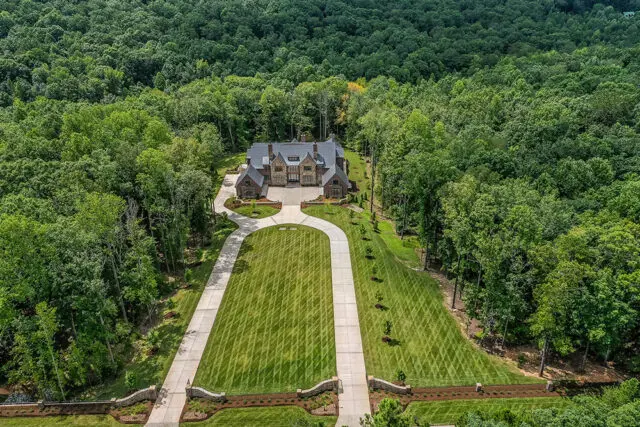 Stone house with manicured lawn and long driveway, surrounded by dense forest.